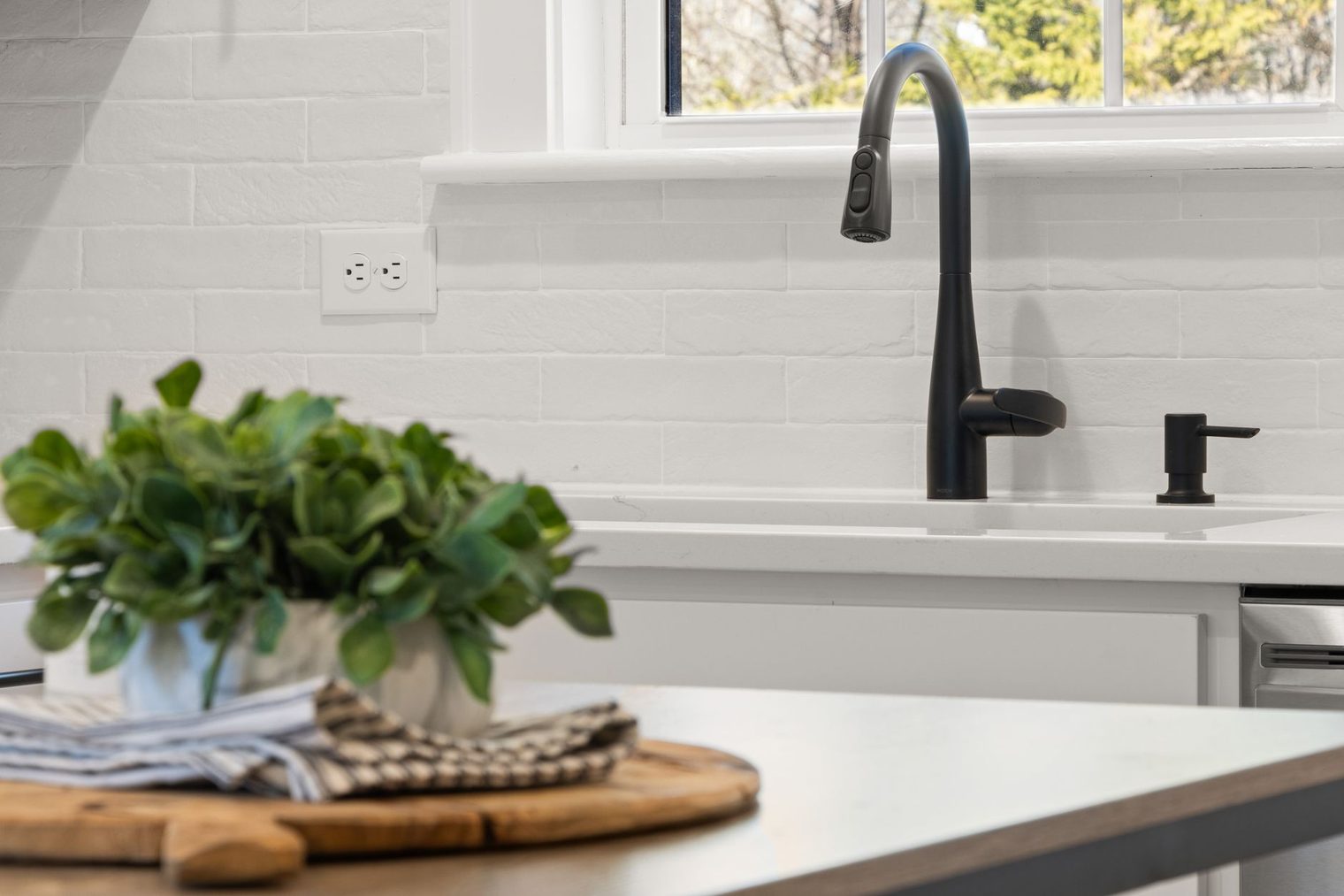 White whitewashed brick backsplash behind a kitchen sink with a black faucet and a potted plant
